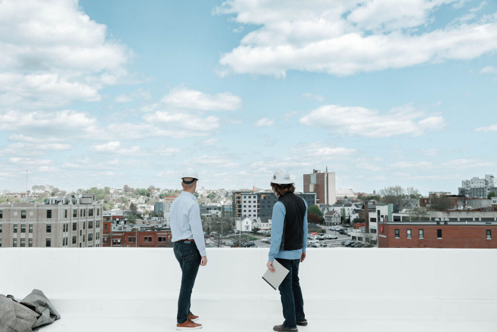 Two men in hard hats, standing on a building's roof and looking out over the city skyline.