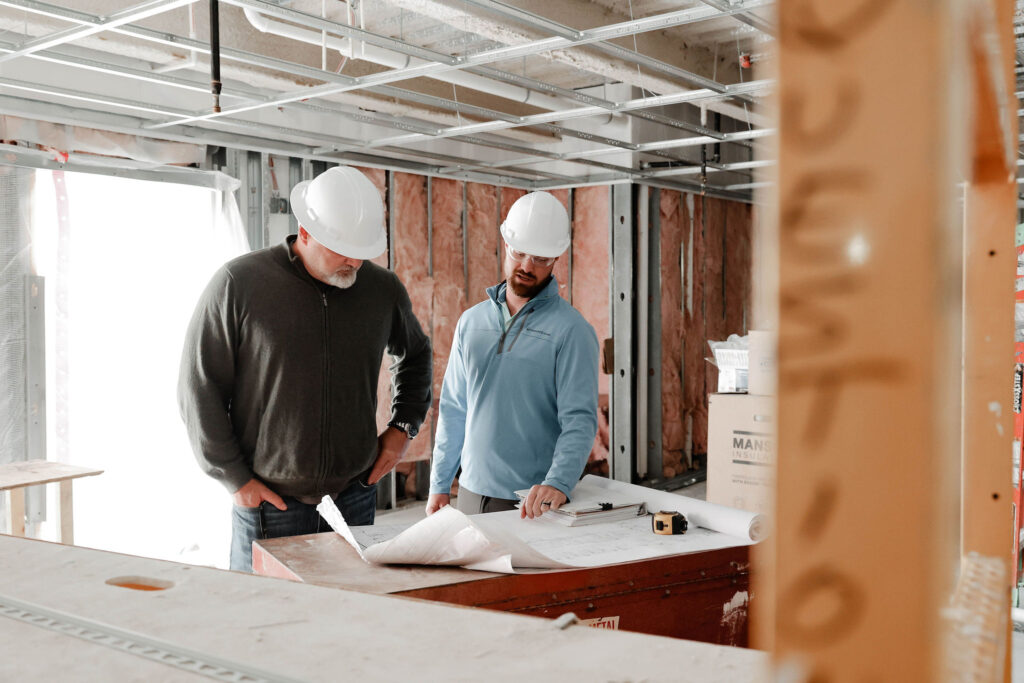 Two men in hard hats looking over building plans on the table in an unfinished room.